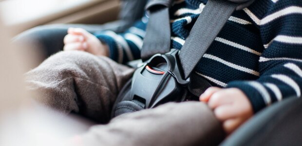 Auf dem Bild ist der Körper eines Kleinkindes zu sehen, welches angeschnallt im Auto sitzt.